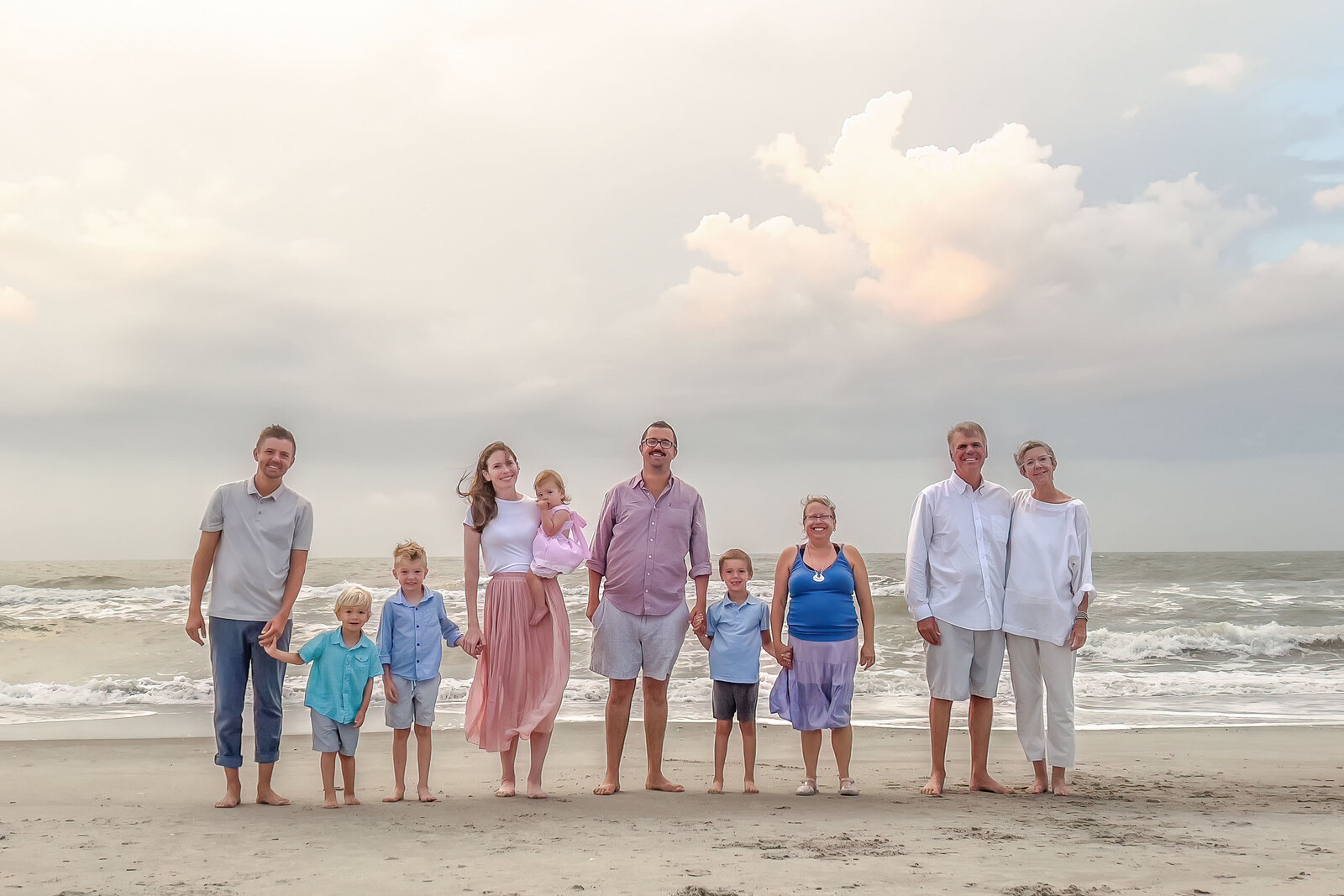 A multi-generational family poses on a windy beach, with dramatic clouds and ocean waves behind them. Dressed in a mix of pastels and whites, they capture a joyful family gathering. Perfect for professional family photography in NC, highlighting dynamic seascapes.