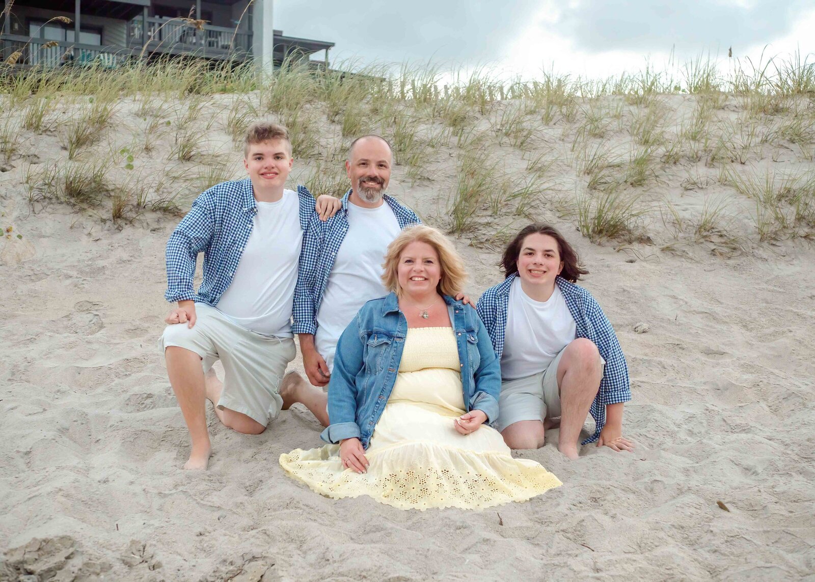 A family of four sits on the sandy dunes of Wrightsville Beach, wearing matching denim and white outfits. The casual pose and beach grass create a cozy atmosphere. This image is ideal for affordable family photography in Wilmington, NC, capturing relaxed family interactions.