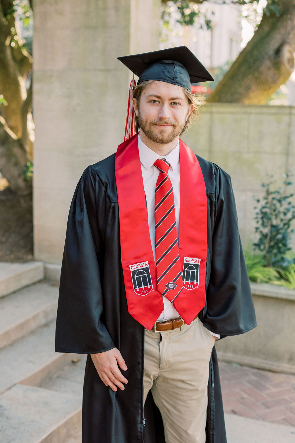 Guy wearing UGA graduation outfit stands under UGA arch