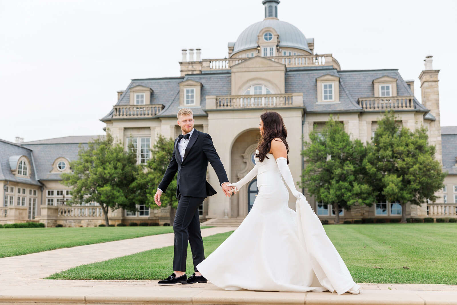 Elegant bride in a strapless white gown and gloves walking hand-in-hand with the groom in a black tuxedo in front of a grand French chateau-style wedding venue in Hickory Creek, Texas, surrounded by manicured lawns and classic architecture.