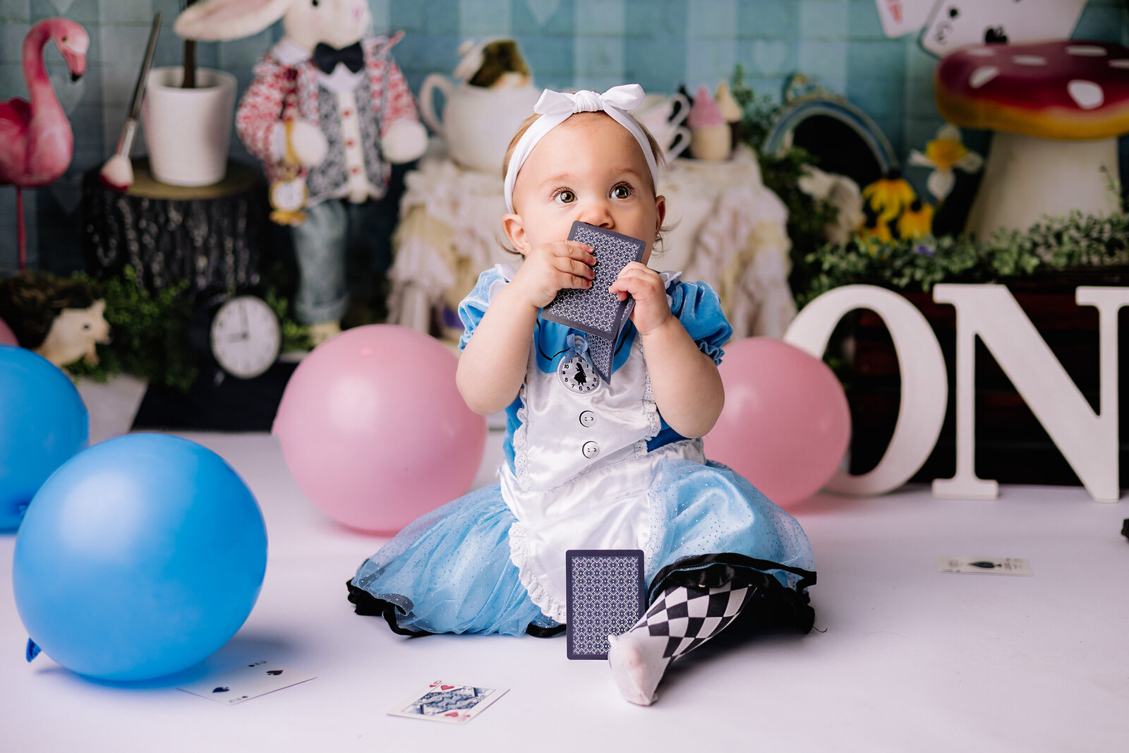 Nibbling on a playing card, a baby girl sits amongst a mess of balloons during her milestone session