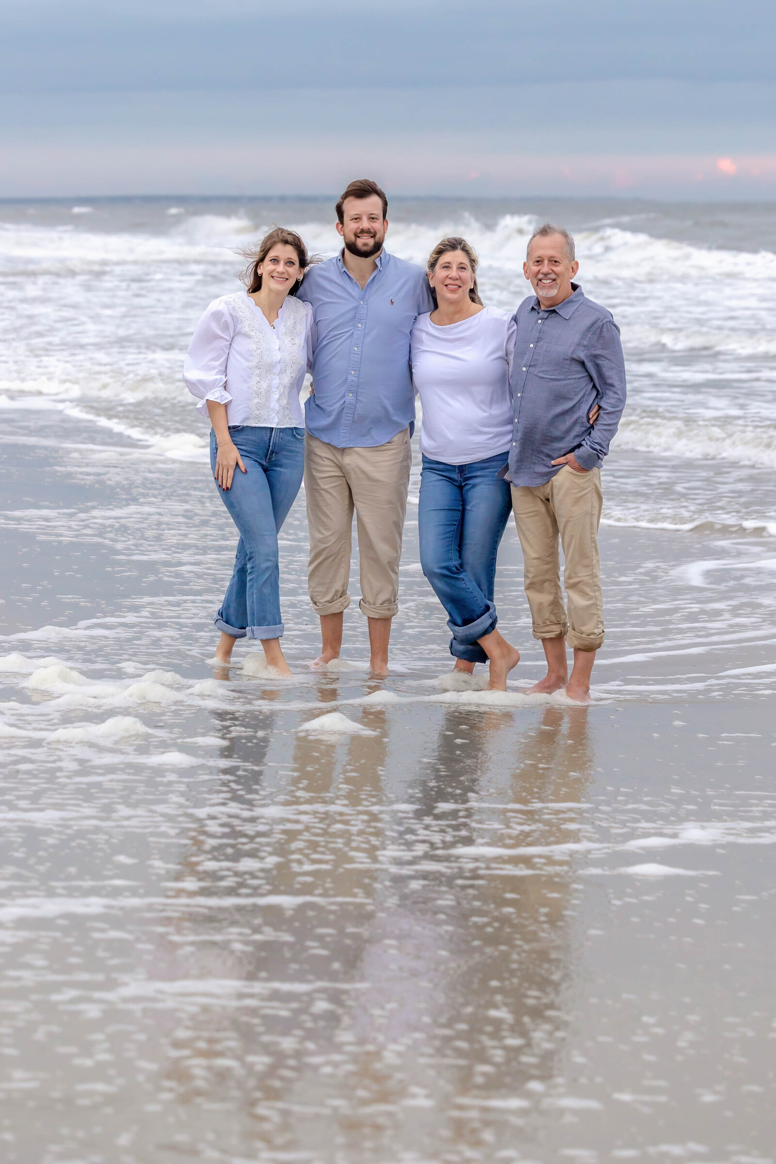 A family of four stands barefoot in the ocean waves, dressed in blue and white attire. The serene seascape and gentle surf create a peaceful setting, ideal for professional family photography in NC, capturing intimate and natural coastal moments.