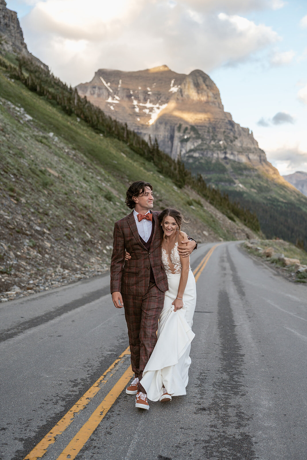 A joyful bride and groom walk arm in arm down a winding mountain road surrounded by rugged cliffs and soft golden light during their Glacier National Park elopement, captured by Sydney Breann Photography.