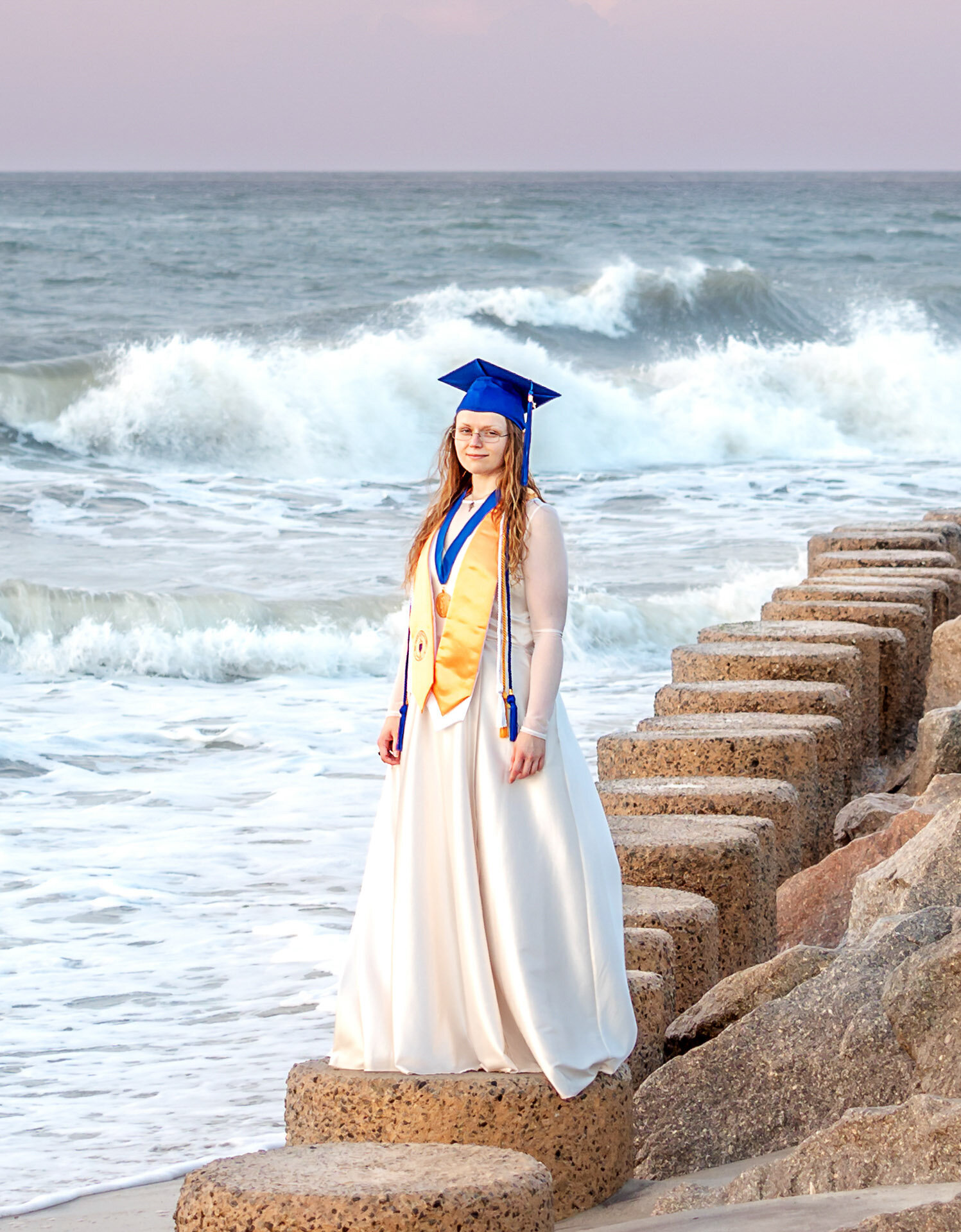 A CFCC college senior in a blue cap and gown standing on beach rocks with waves crashing behind in Wilmington, North Carolina. This striking image highlights the excitement of graduation, perfect for artistic senior photography by the sea.