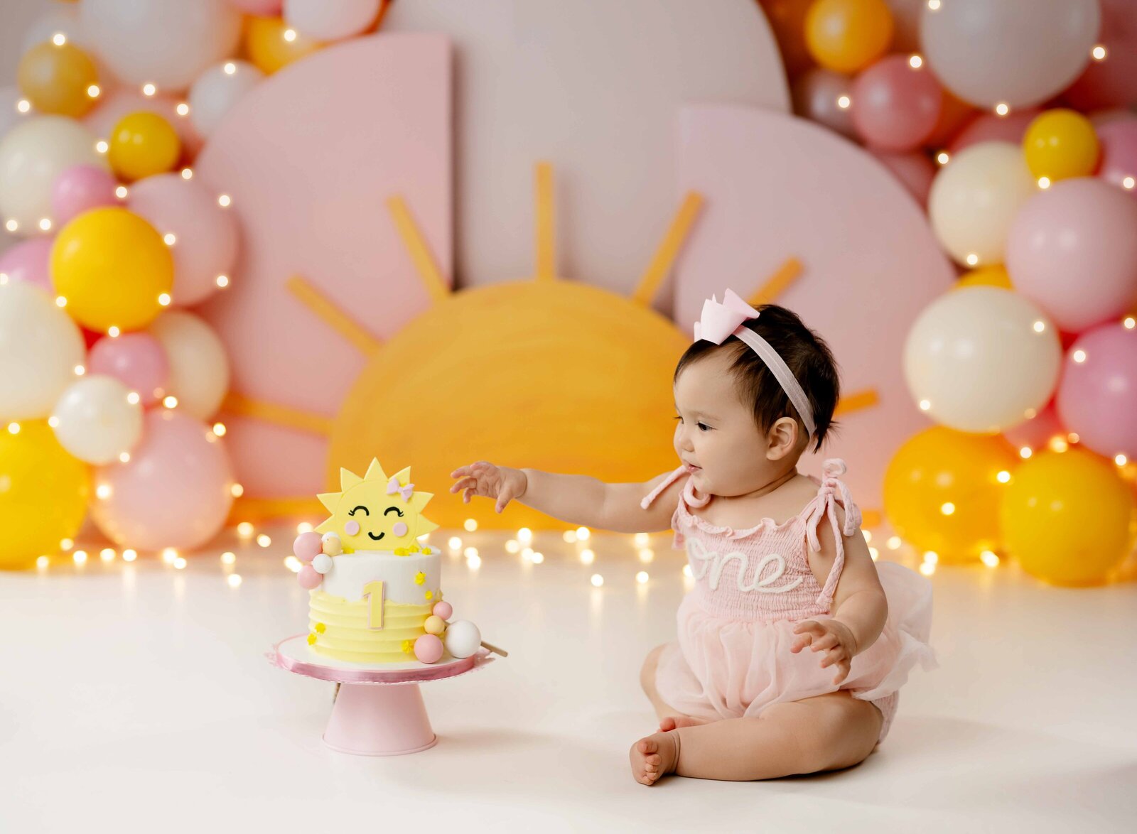 Baby reaching to touch the sunshine cake. Pink and yellow balloons in the backdrop with sun and arches. 