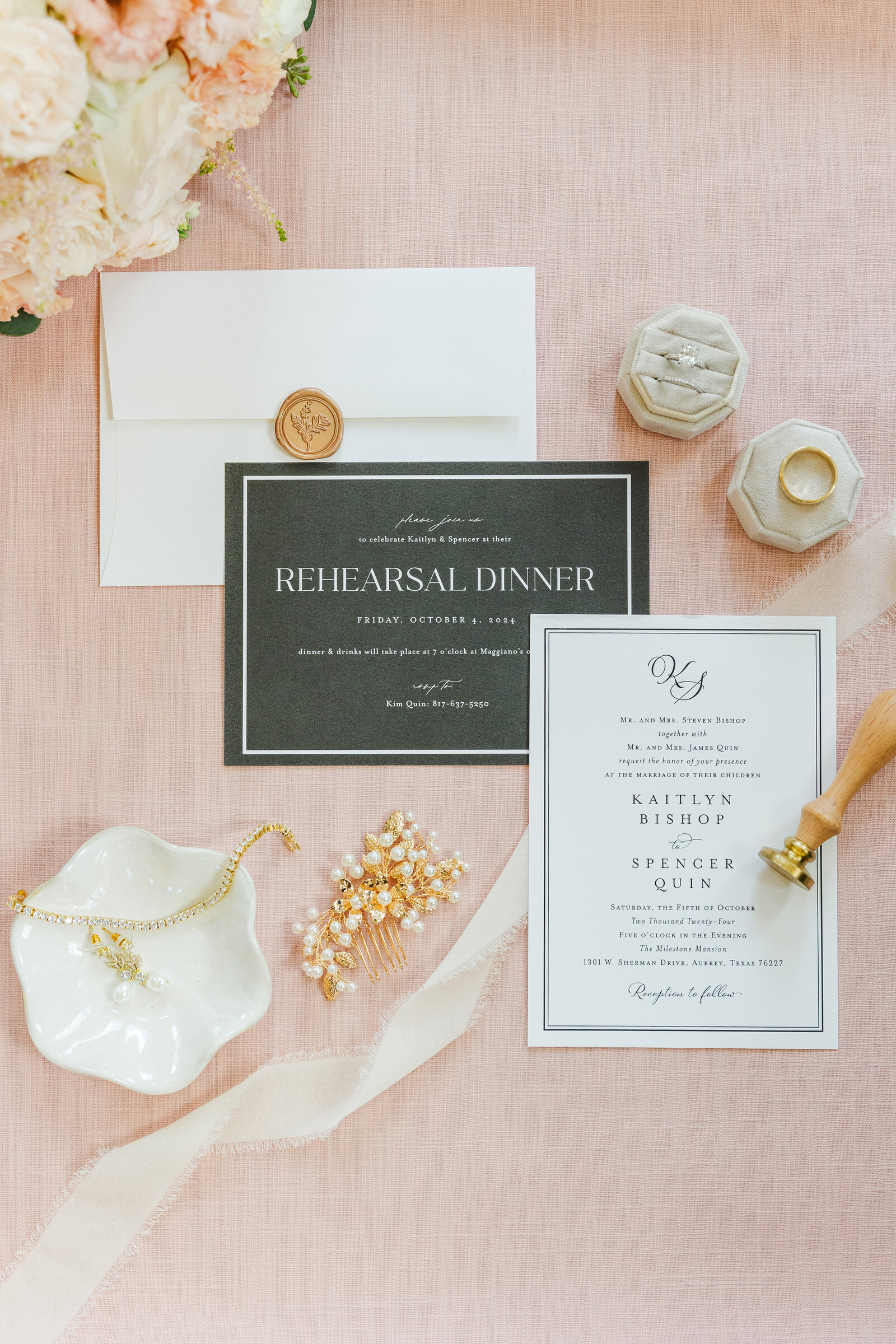Flat lay of black and white rehearsal dinner and wedding invitations with blush velvet ring boxes, gold accessories, a wax-sealed envelope, and soft floral accents styled on a blush pink linen background.
