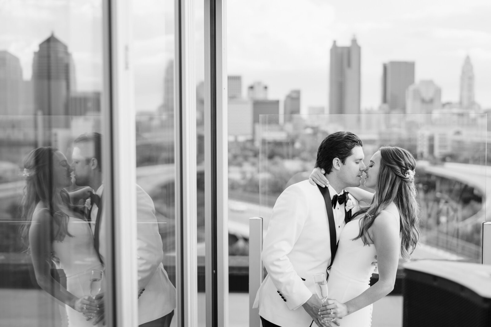 A groom in a white tuxedo dips his bride and kisses her with the Columbus skyline in the background