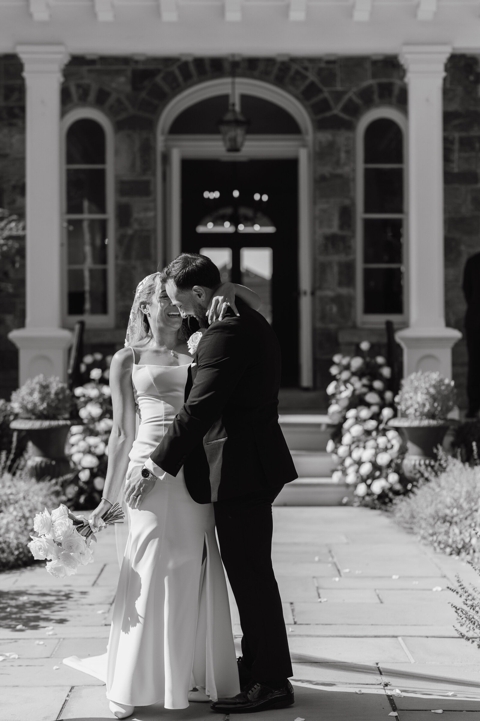 Black and white image of bride and groom outside of their wedding venue in simple luxury white wedding gown and black tuxedo | Emma LeeAnn Photography