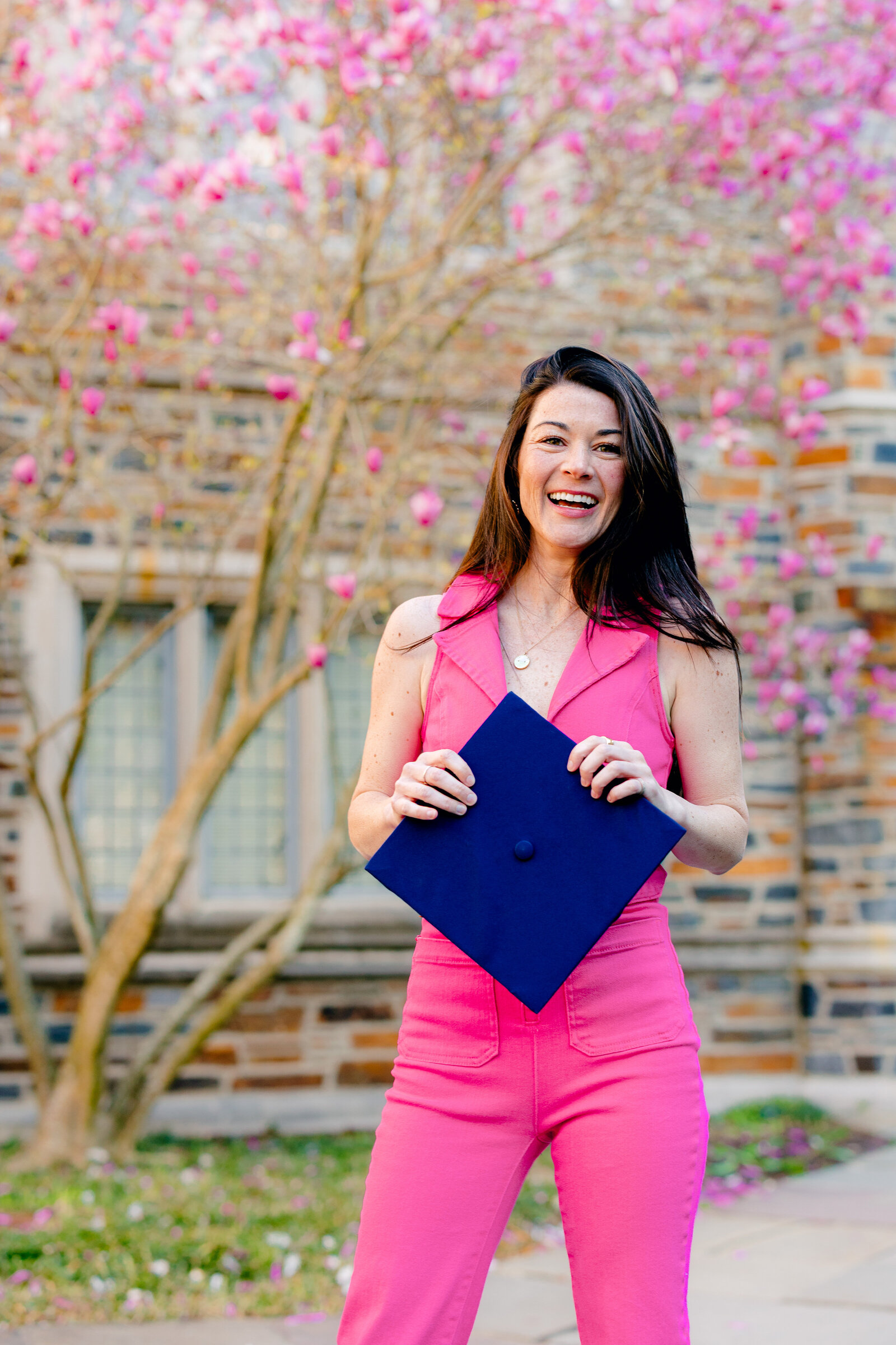 Duke University graduate wearing a pink jumpsuit holding her navy cap and smiling during spring graduation portraits.