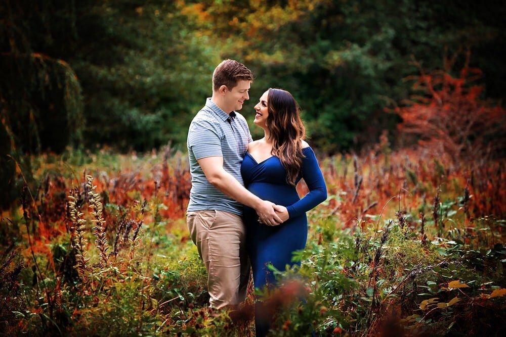 Pregnant couple smiling at eachother in fall field.