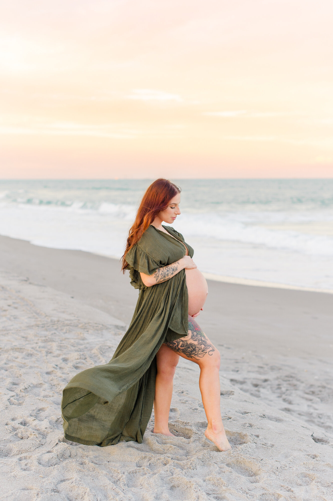 Beautiful expectant mother stands on the beach holding her belly at sunset during her maternity session on Cocoa Beach east of Orlando