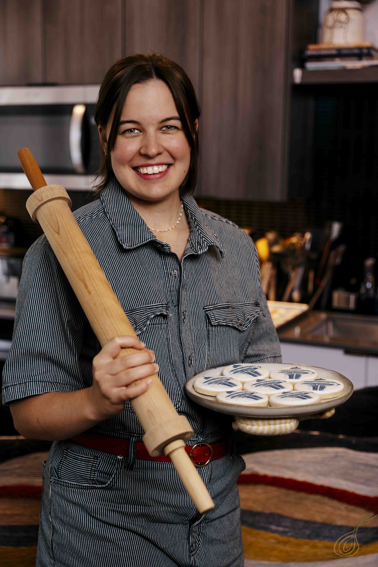 A woman stands confidently, holding a rolling pin in one hand and a beautifully decorated cookies in the other.