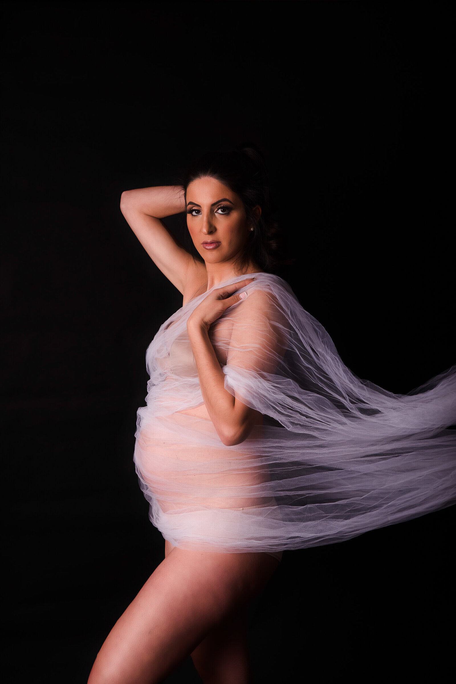 A mother-to-be poses confidently, wrapped in flowing sheer white fabric, looking directly into the camera against a dark backdrop.