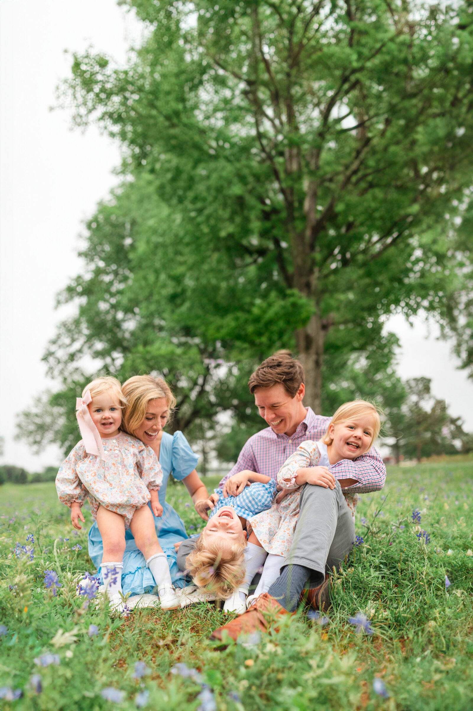 happy family of 5 with three under 5 in a beautiful green outdoors field. Family is under a tree all laughing while they tickle each other in joy
