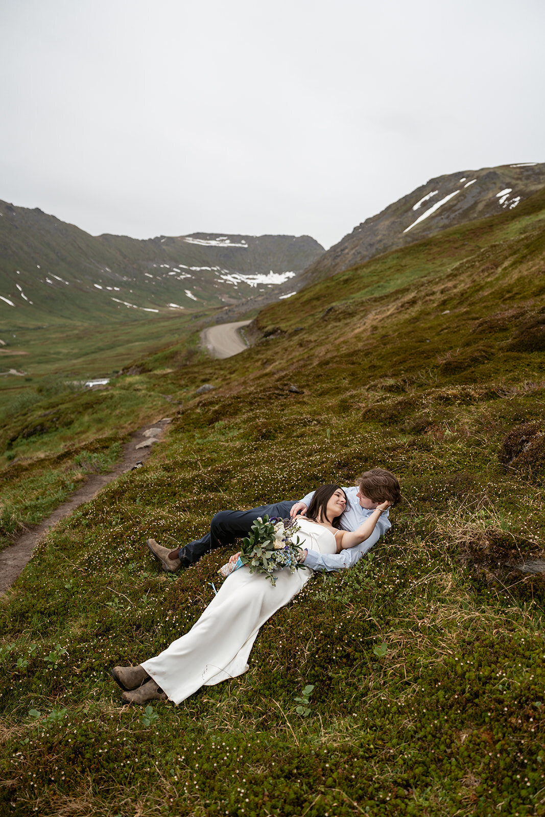 A couple lies together on a mossy hillside surrounded by misty mountains and patches of snow during their Alaska elopement, captured by Sydney Breann Photography.