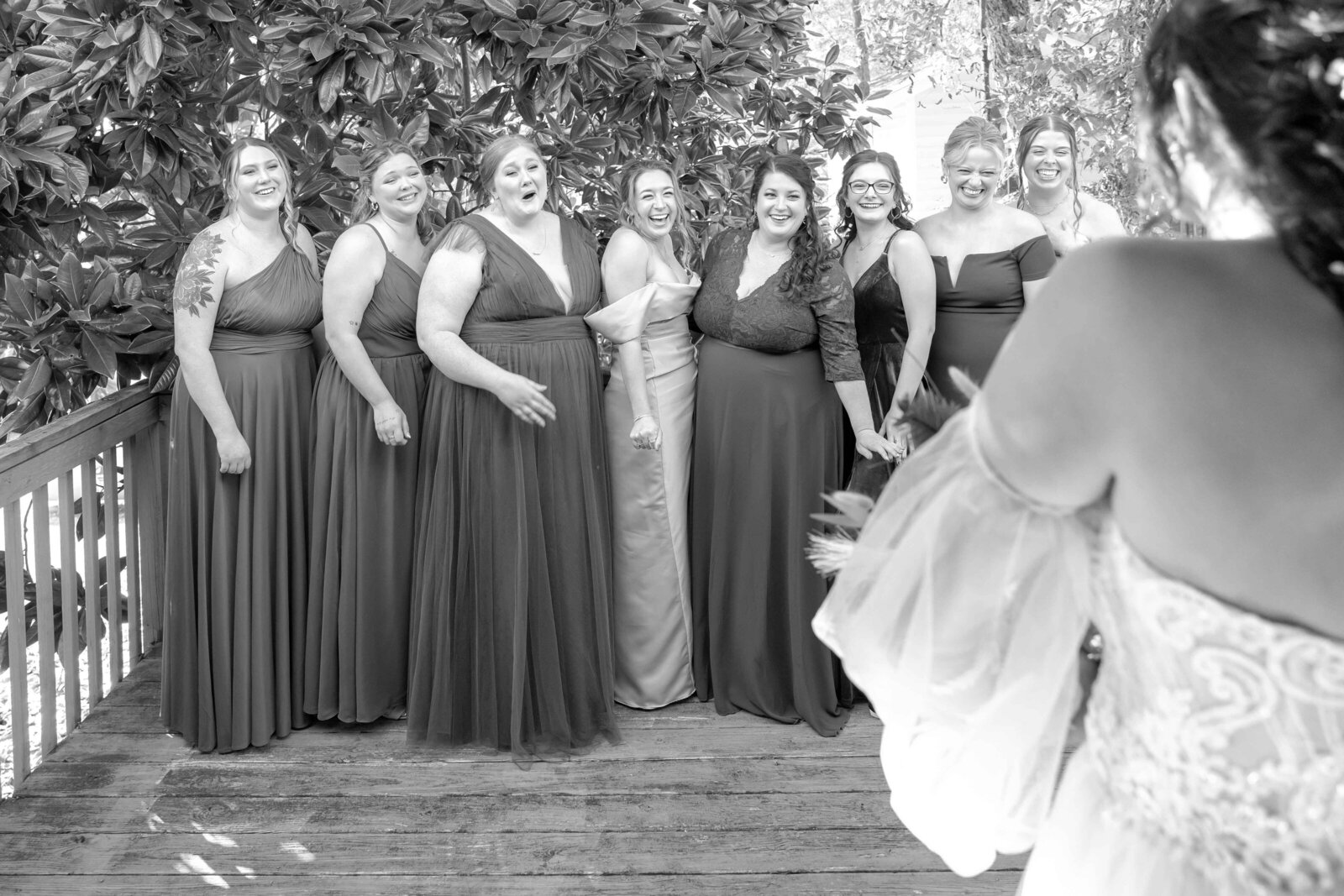 A group of bridesmaids reacts with excitement during a first look with the bride, captured in black and white on a wooden deck during a Wilmington, NC wedding, showcasing authentic and joyful moments.
