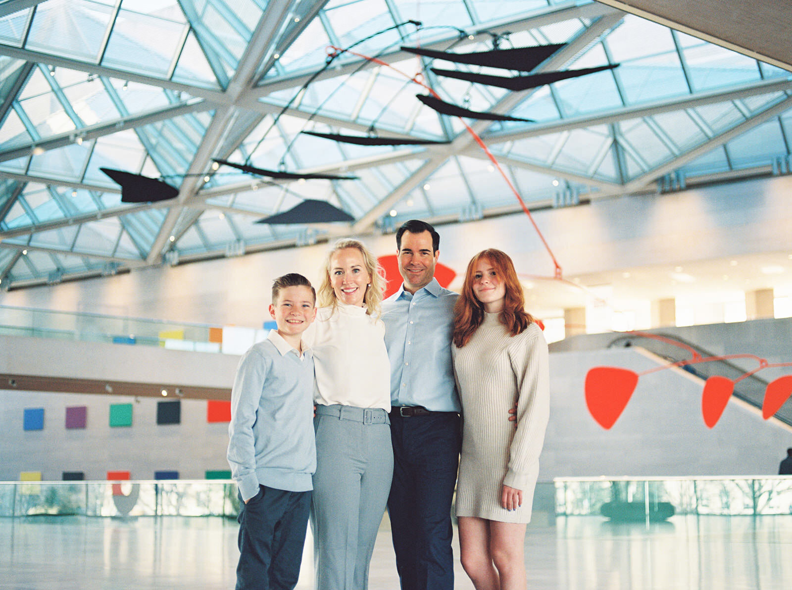 "A stylish family portrait in a modern art gallery, featuring parents with their two children under a bright geometric ceiling with contemporary sculptures in the background."