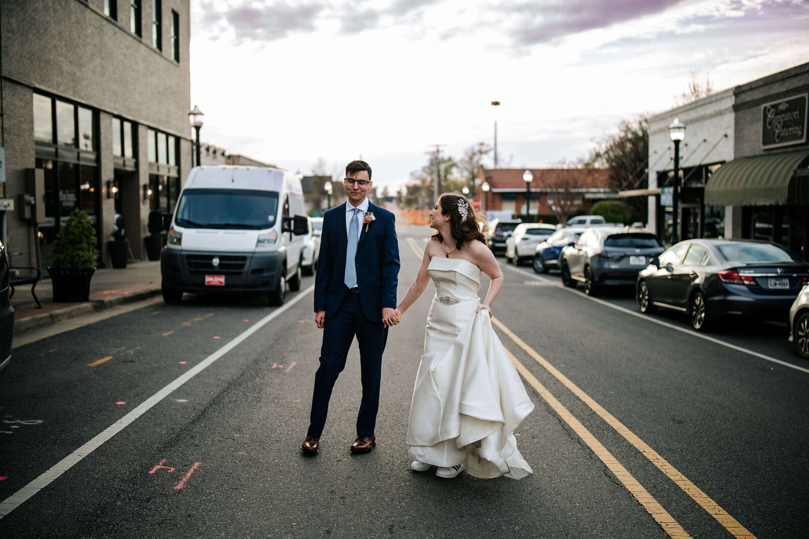 A bride in a white dress and a groom in a blue suit stand holding hands in the middle of a city street, with parked cars and buildings in the background under a cloudy sky.