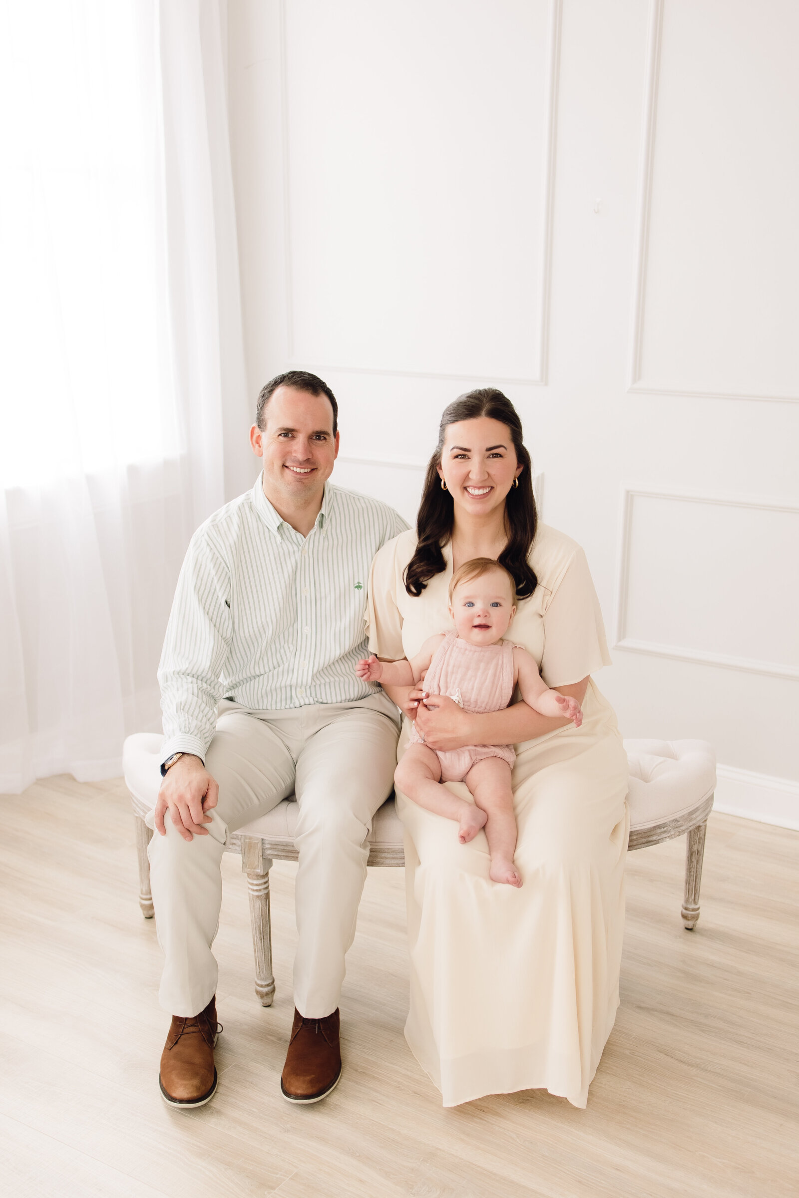 Family of three in light neutral clothing sitting on a bench in a bright studio