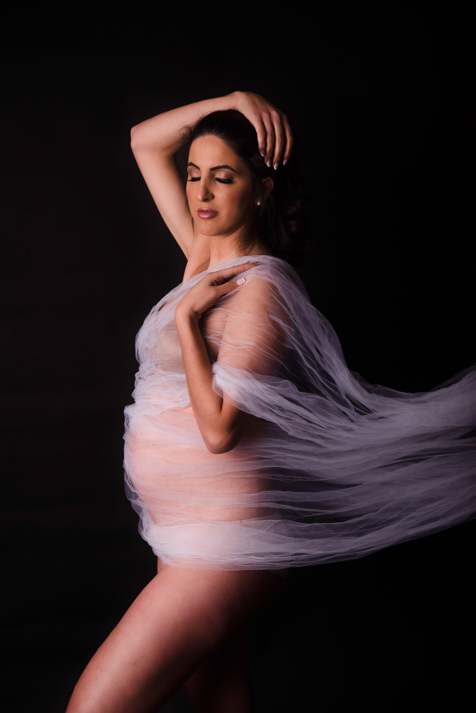 A glowing expectant mother is wrapped in soft white sheer fabric, creating a dreamy, angelic silhouette against a dark background.