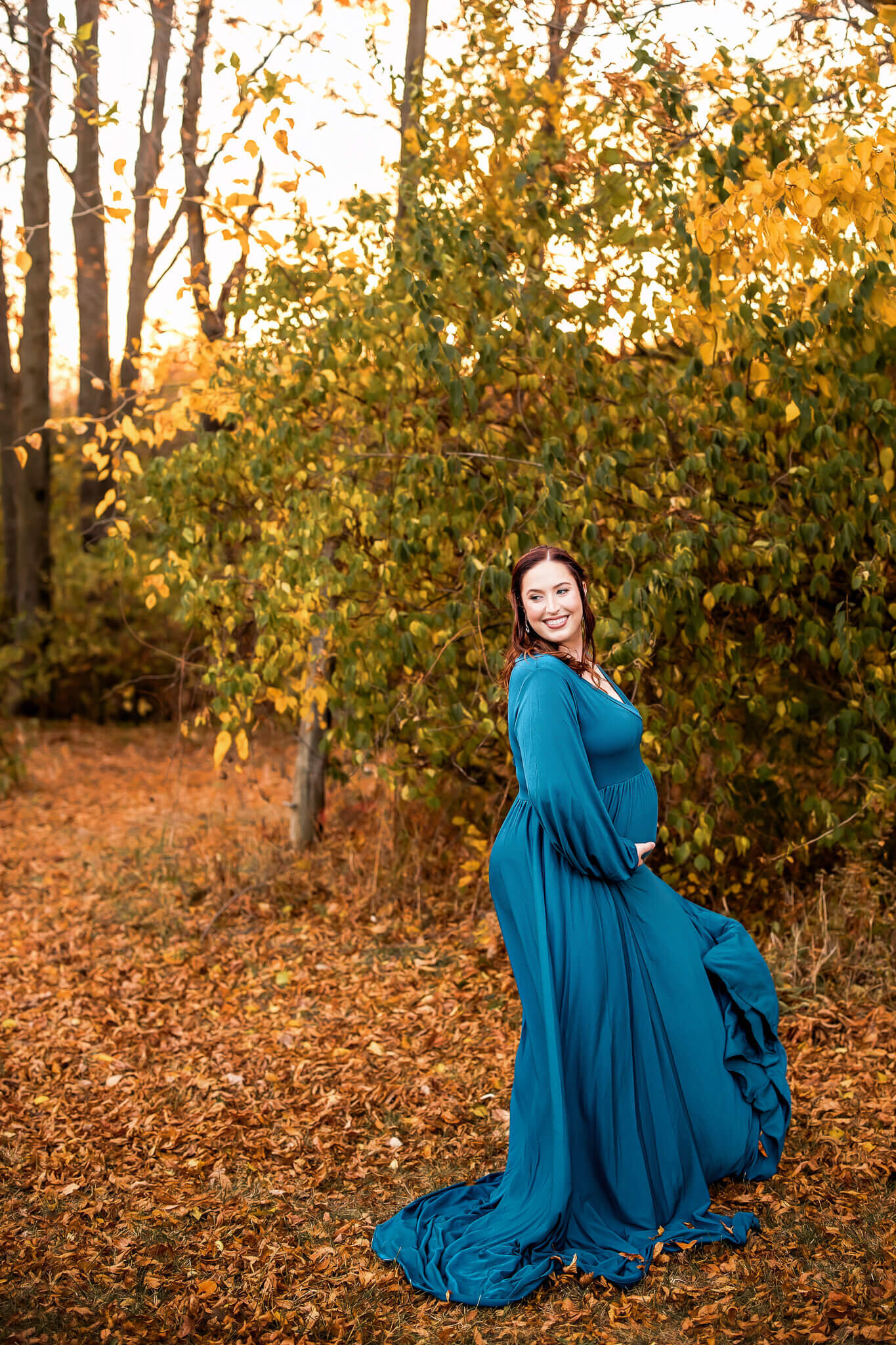 A woman in a long, flowing teal dress stands on fallen autumn leaves, smiling and holding her dress in a wooded area with golden foliage—perfect for stunning maternity portraits by a Bloomington maternity photographer.