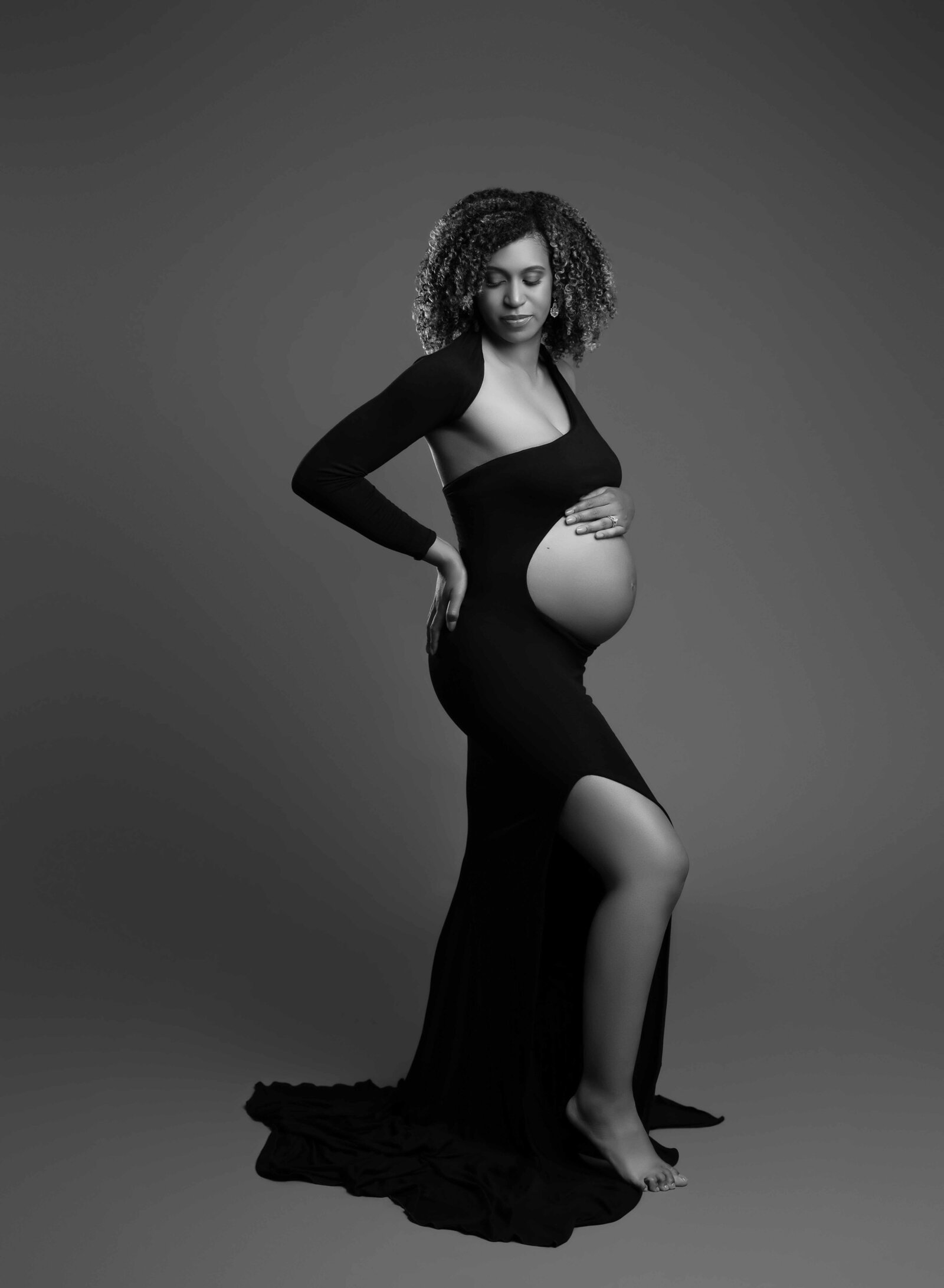 Pregnant women in elegant black maternity gown with hand on hip and other hand on belly. Photo taken at Atlanta Maternity photo studio in Woodstock GA.