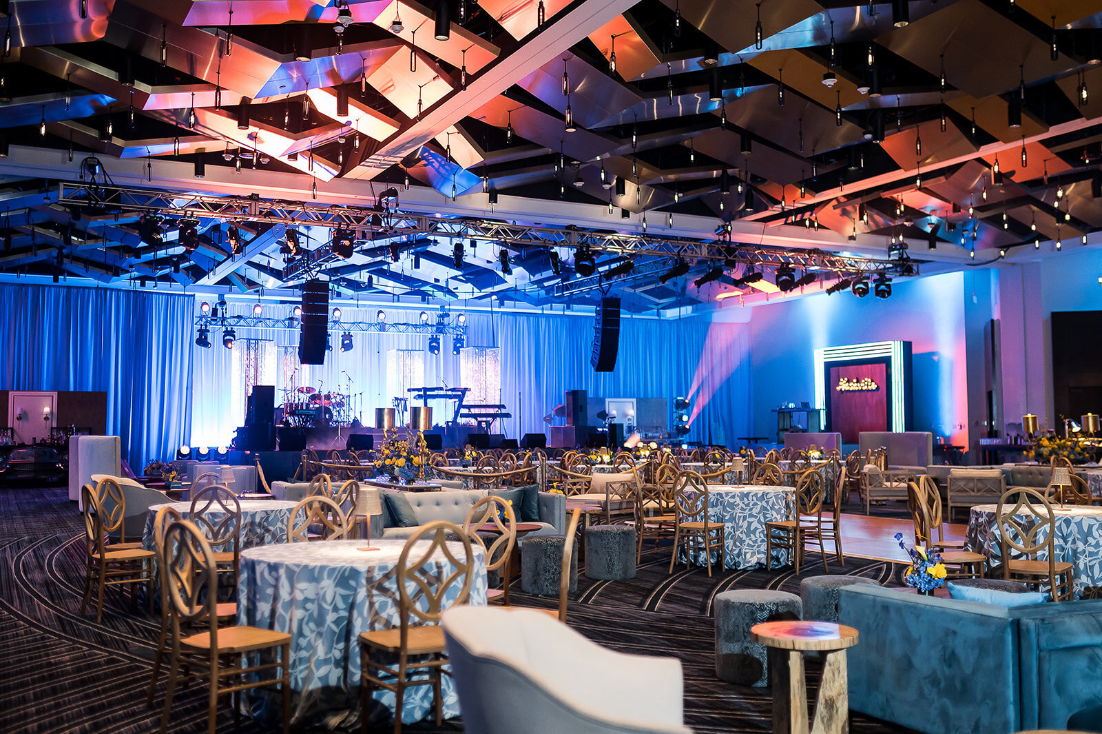 Modern corporate event lounge in Nashville, TN with dramatic stage backdrop, ambient blue and magenta uplighting, round tables with patterned linens, wooden chairs, and plush seating, styled by Wells Design Co for a high-end business entertainment experience.