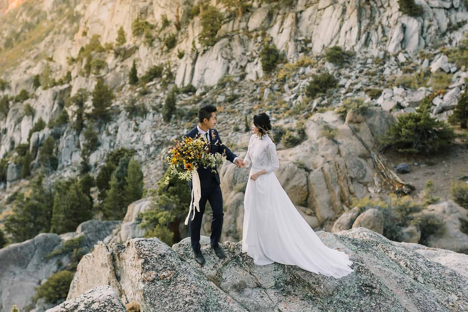 A very adventurous bride and groom climb up the side of a rock face overlooking a beautiful mountain valley on their mountain wedding day. 