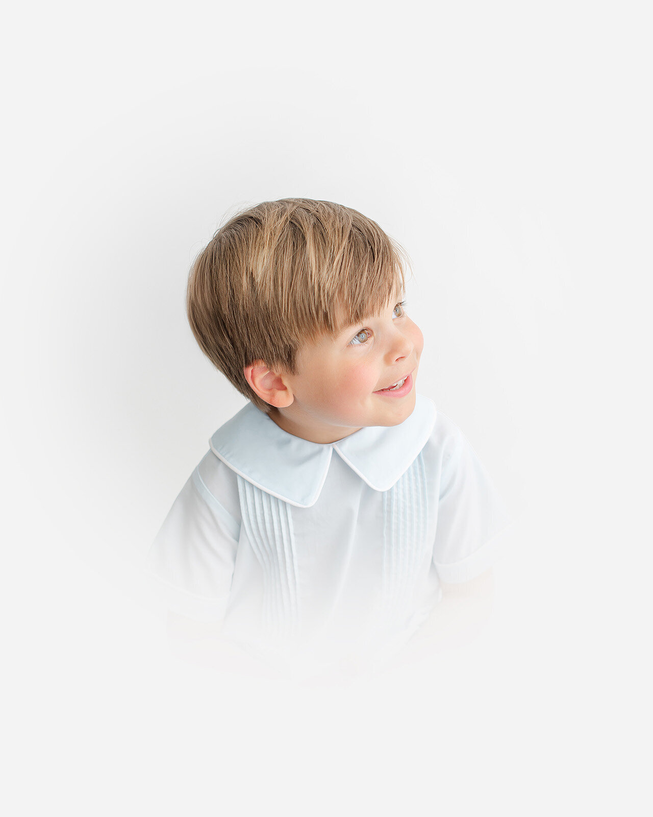 A young child with light brown hair smiles and looks upward, embodying the timeless charm of heirloom photography, wearing a white shirt with a wide collar against a soft white background.