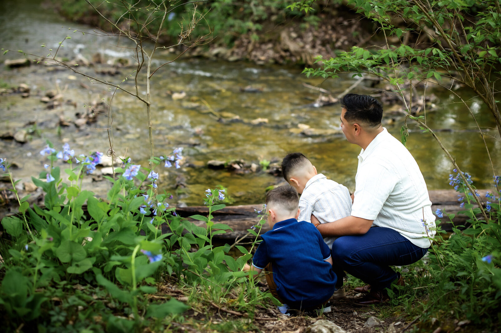 A man and two young boys crouch by a stream in a lush, green forest surrounded by wildflowers, looking at the water together—an outdoor portrait captured by a Bloomington family photographer.
