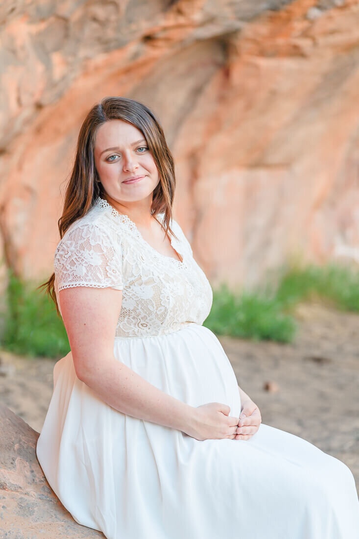 A pregnant woman wearing a long white maternity dress sits on a rock at Red Ledges Picnic Area in Spanish Fork Canyon. Captured by Melissa Woodruff Photography