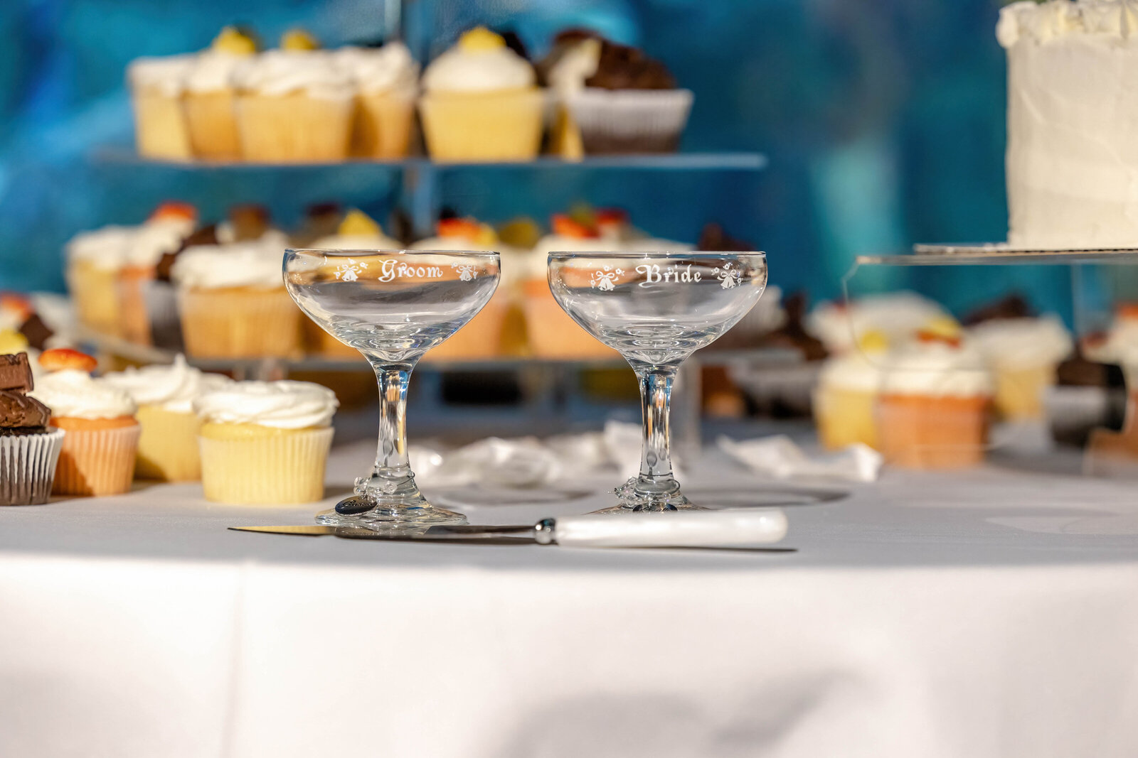 Bride and groom champagne glasses engraved with "Bride" and "Groom" on a dessert table filled with cupcakes and a wedding cake, captured during a Pine Knoll Shores, NC wedding, blending elegance and sweetness.