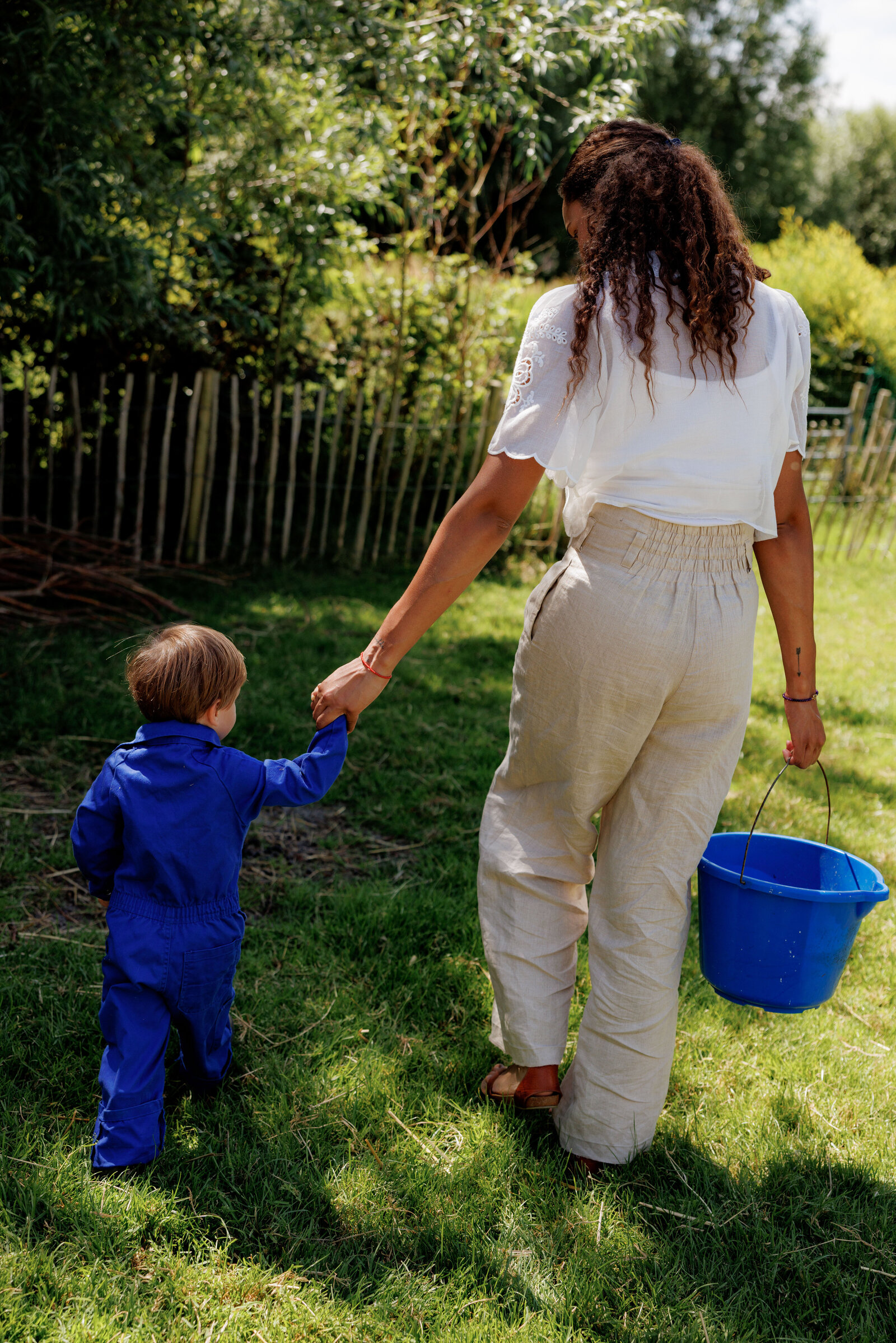 Volwassene houdt de hand van een kind in een blauwe overall vast terwijl ze samen door het gras lopen bij Kinderdagverblijf Bij Ons Op De Boerderij.