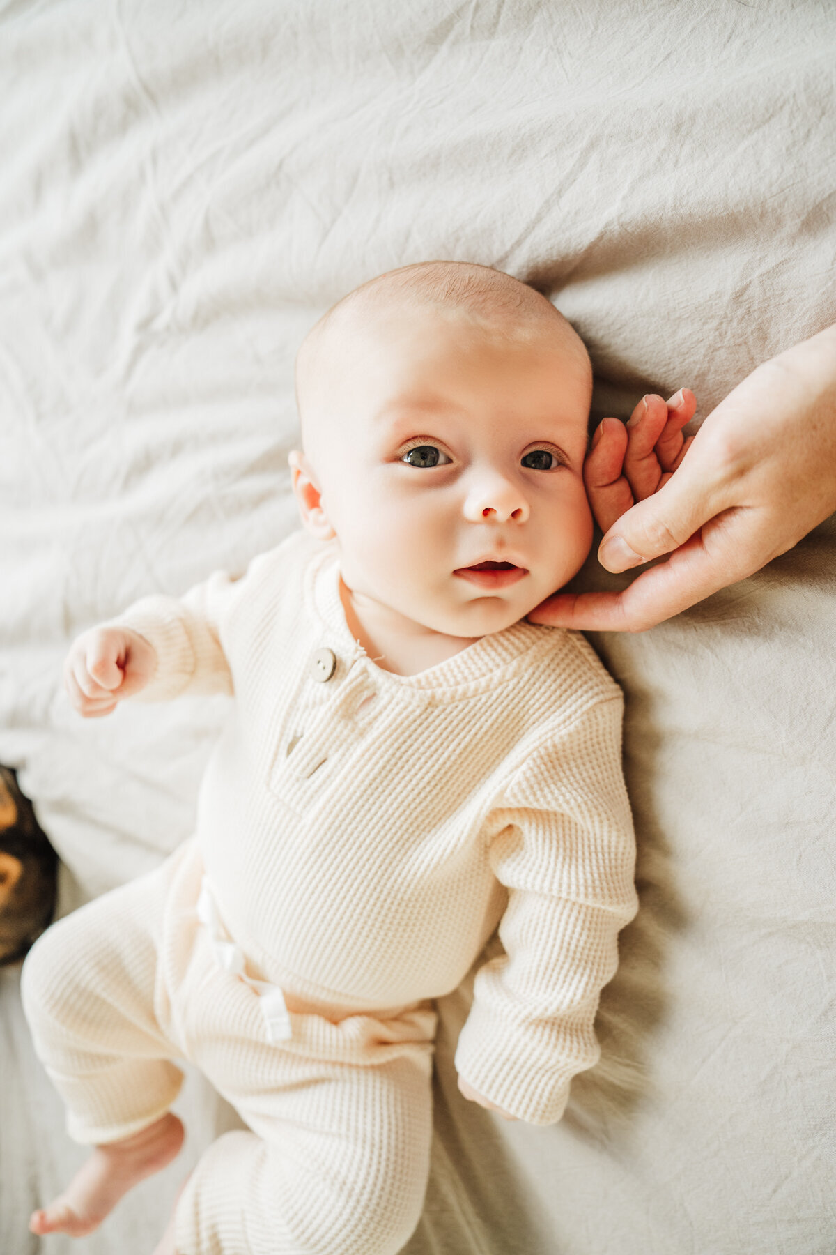 baby boy in cream outfit lays on bed and looks at camera while parents hand strokes cheek