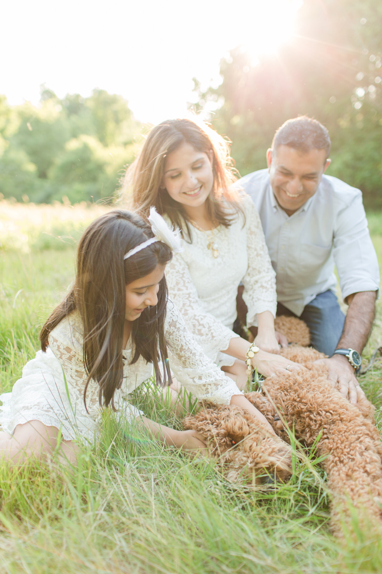 A family of 3 with their dog are sitting in a field of tall grass while petting their dog's curly fur