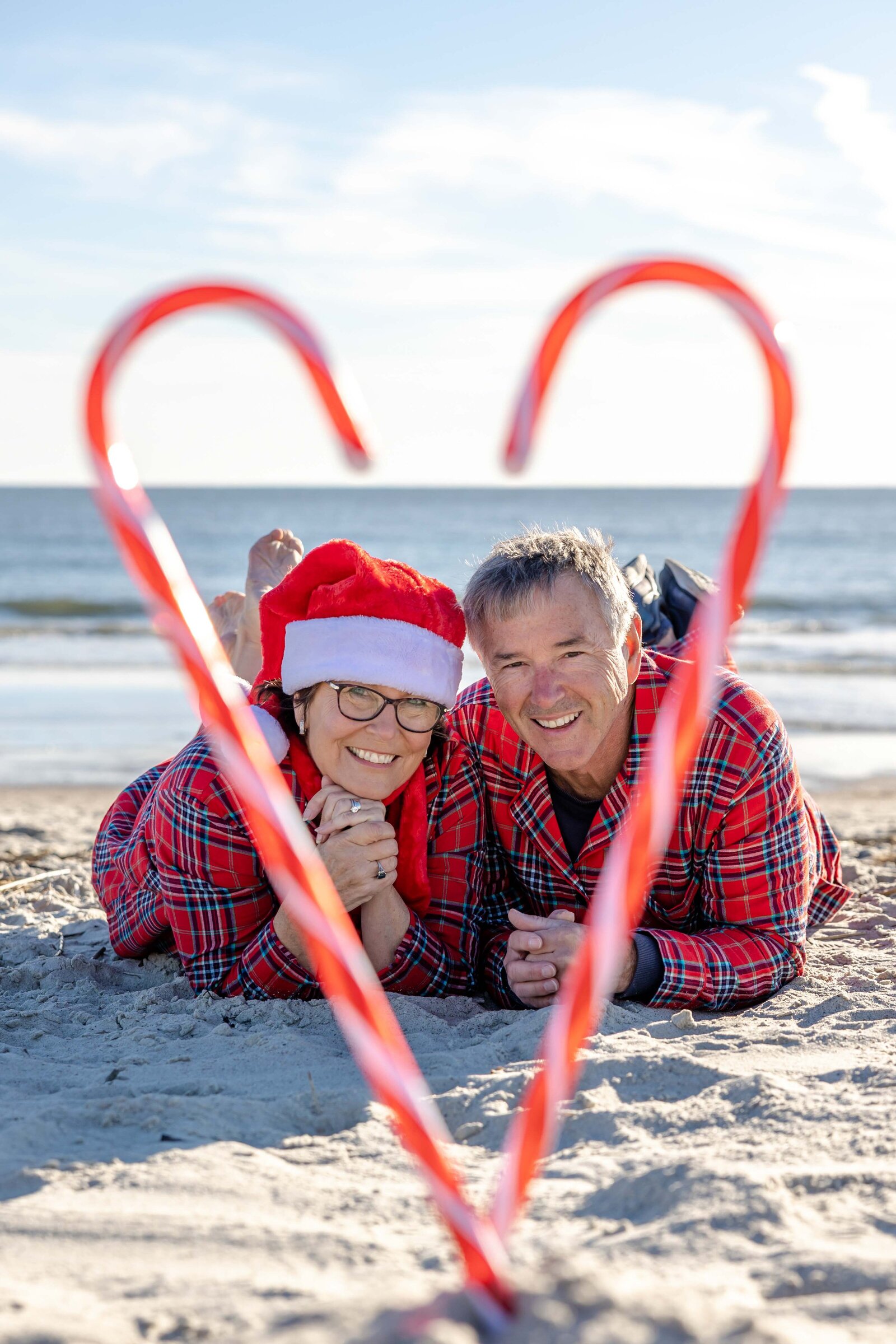 A couple lies on the beach, framed by a candy cane heart. Wearing matching plaid pajamas and a Santa hat, they share a joyful holiday moment by the ocean. Ideal for professional family photography in NC, highlighting festive beachside romance.