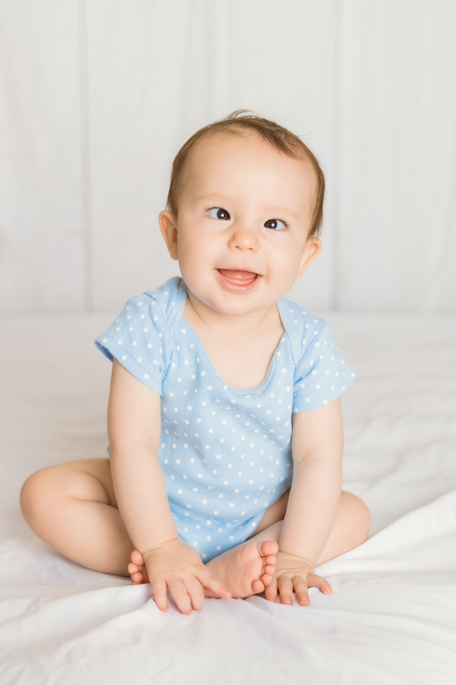 Milestone Photographer, a baby wears a onesie and sits up on the bed sheets smiling