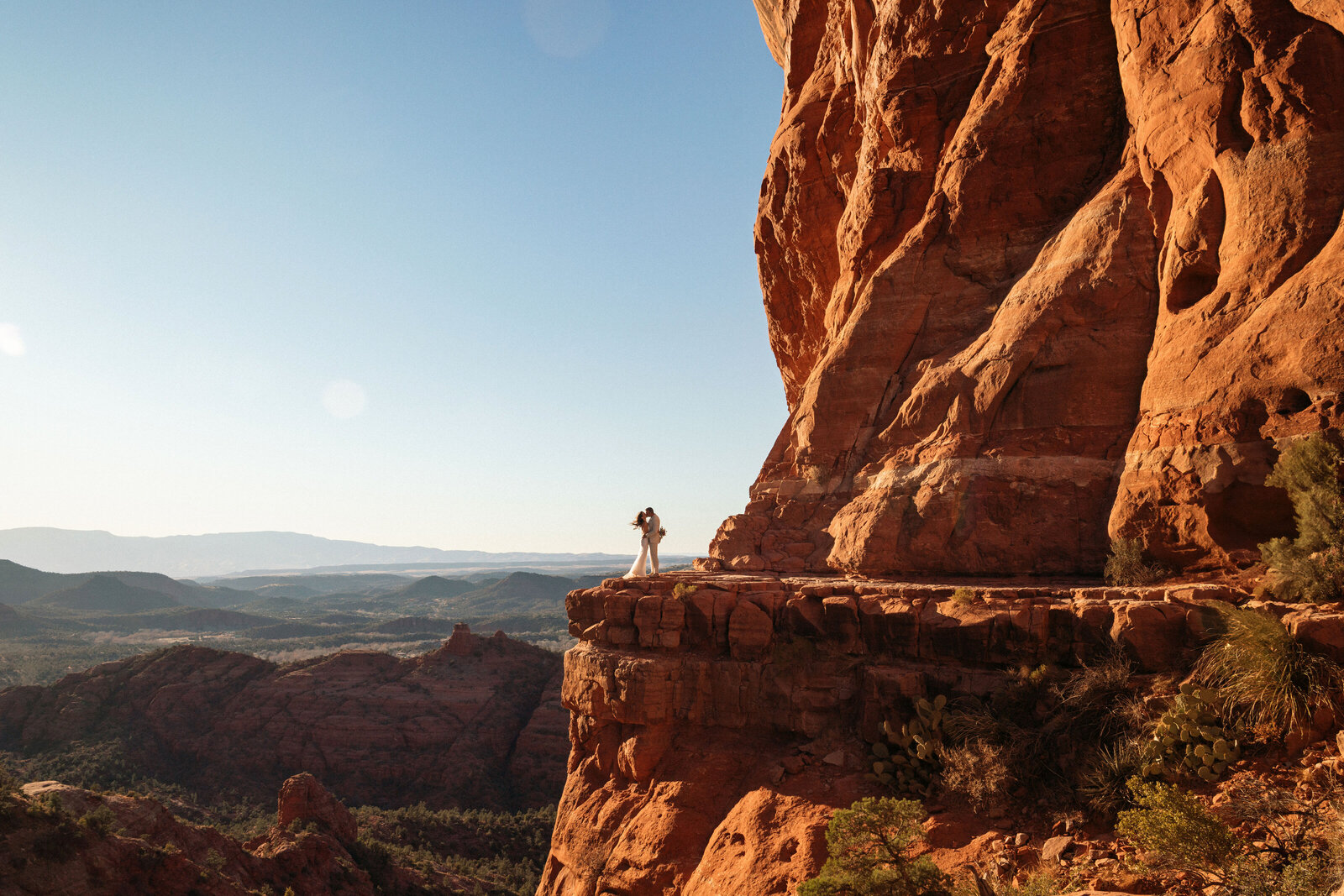 Bride and groom standing on a dramatic red rock ledge at Cathedral Rock in Sedona, Arizona, sharing an intimate moment with sweeping desert views and golden sunlight illuminating the sandstone cliffs.