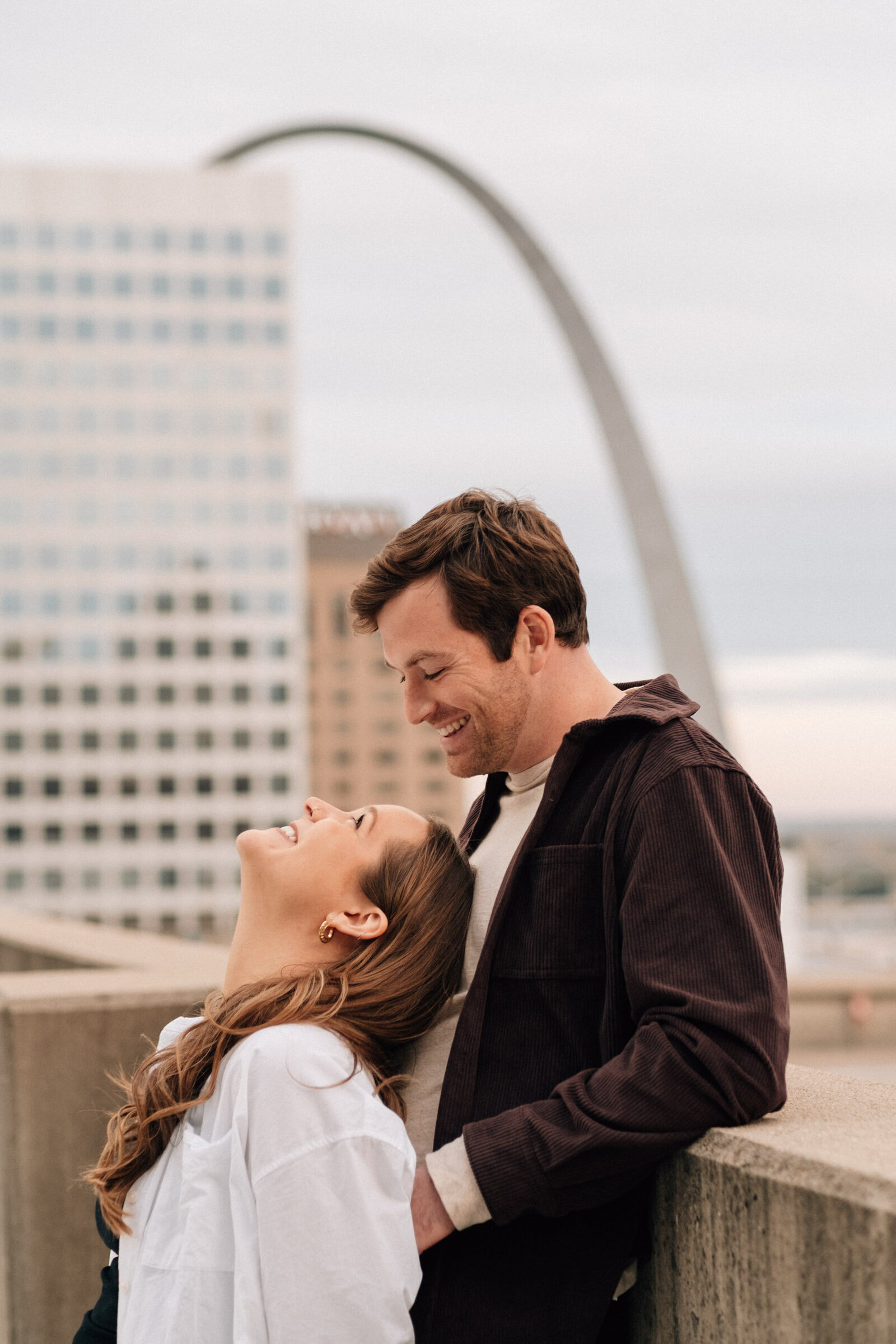 A couple smiles at each other during a rooftop engagement photoshoot in St. Louis, Missouri, with the Arch in the background.