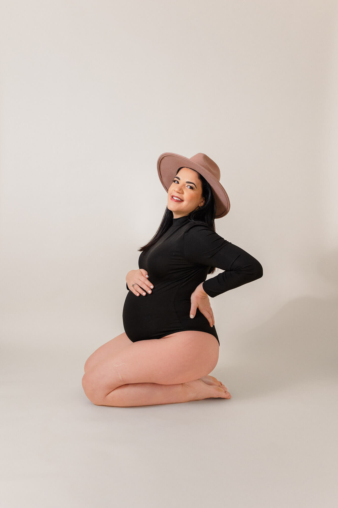 A pregnant woman in a black bodysuit and pink hat posing and smiling at the camera in a Gainesville, VA studio.