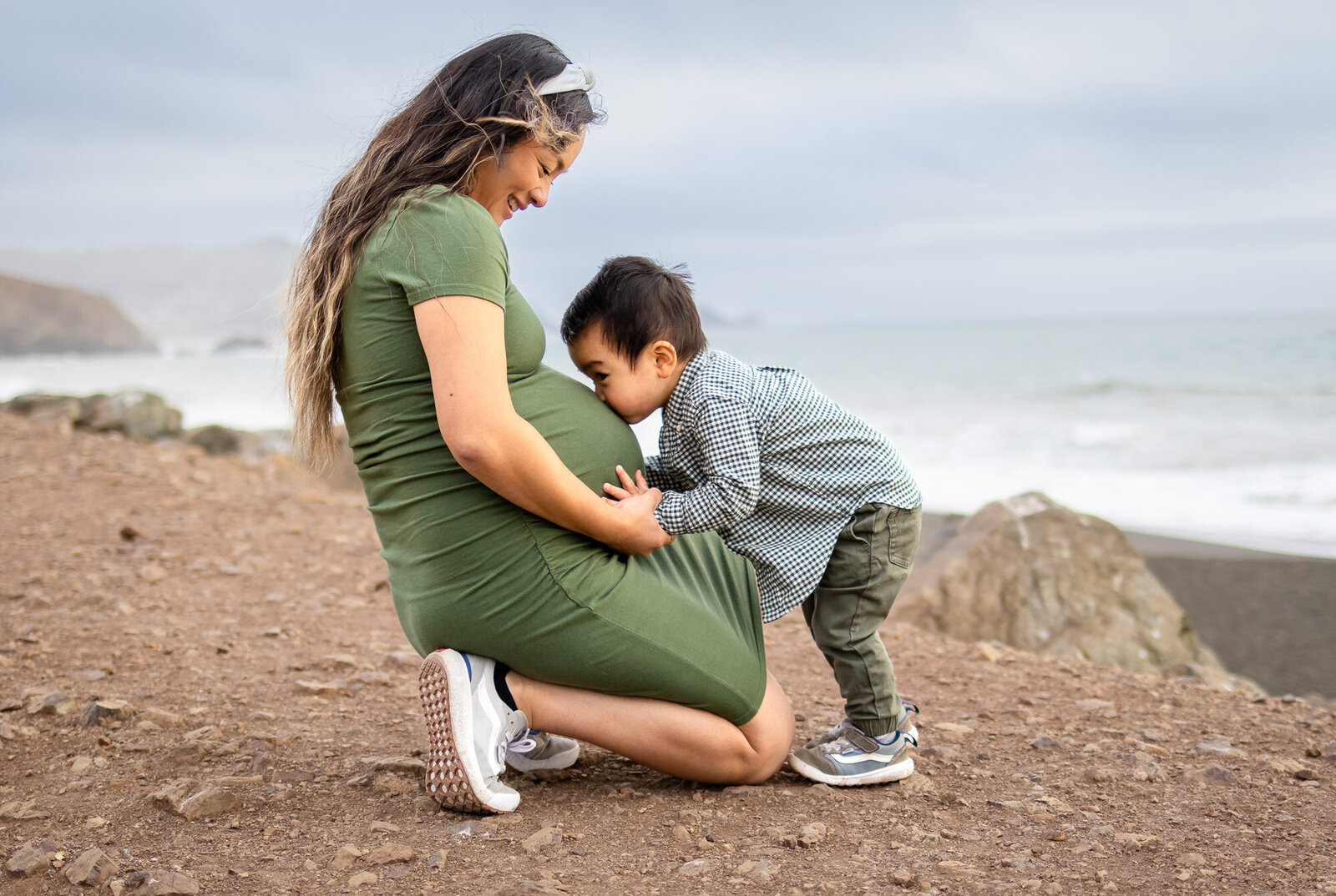 Toddler kissing mom’s pregnant belly while kneeling on a dirt path by the ocean, Bay Area Maternity Portfolio - Ellobelle Photography