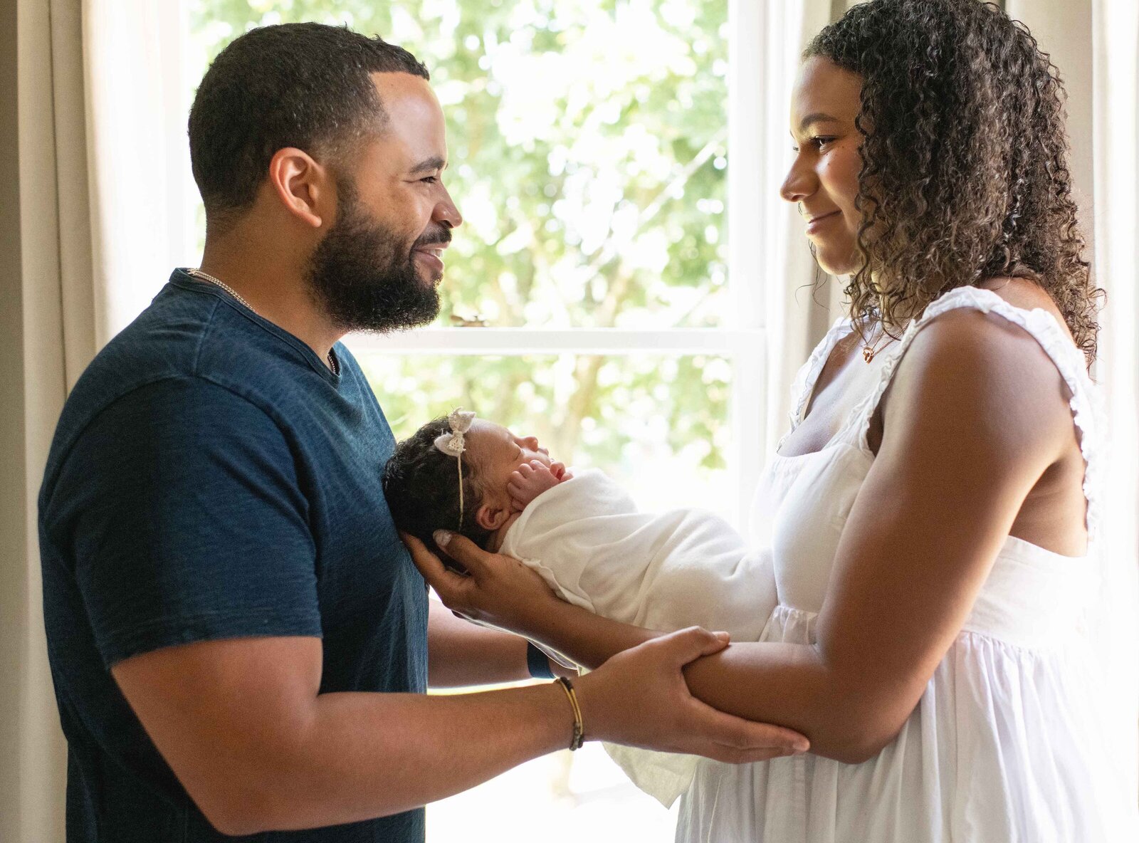 mother and father of a newborn baby standing in front of a window looking at eachother