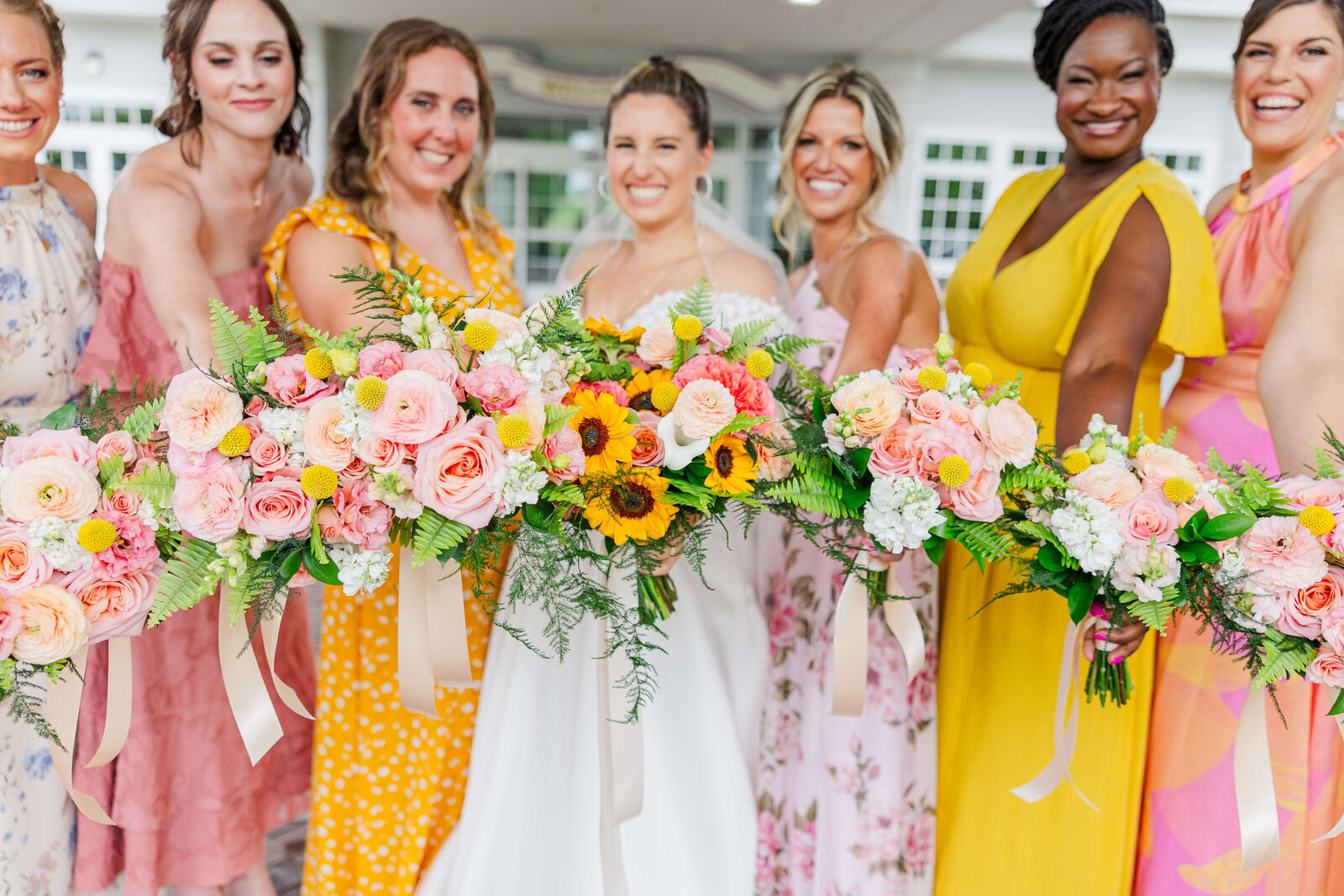 Bridesmaids in bright yellow and pink dresses hold up their bright bouquets with the bride. They are all smiling
