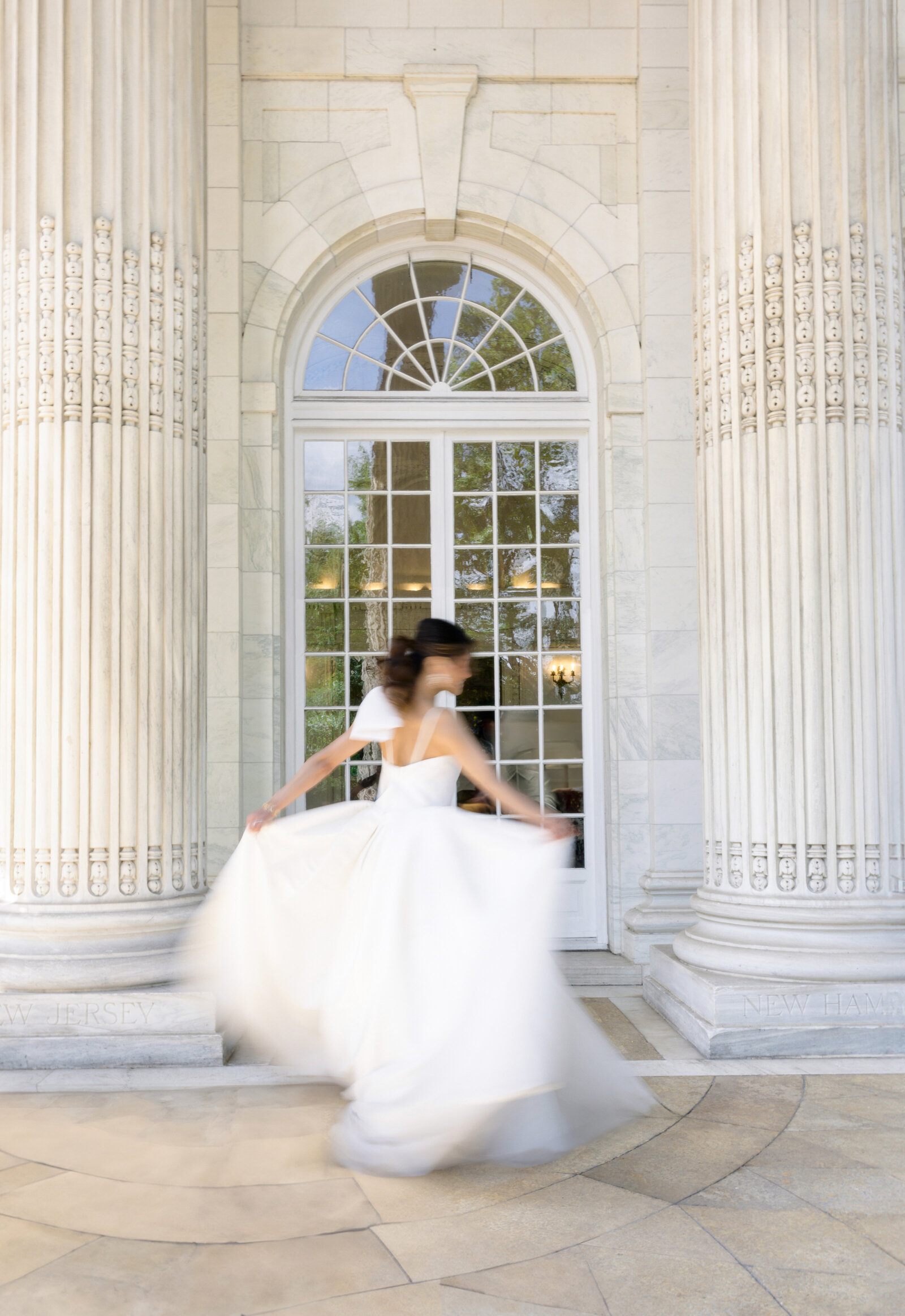 A bride in a flowing white wedding dress twirls joyfully in front of a large arched window. The background features elegant, tall columns and stone architecture, creating a timeless and romantic atmosphere.