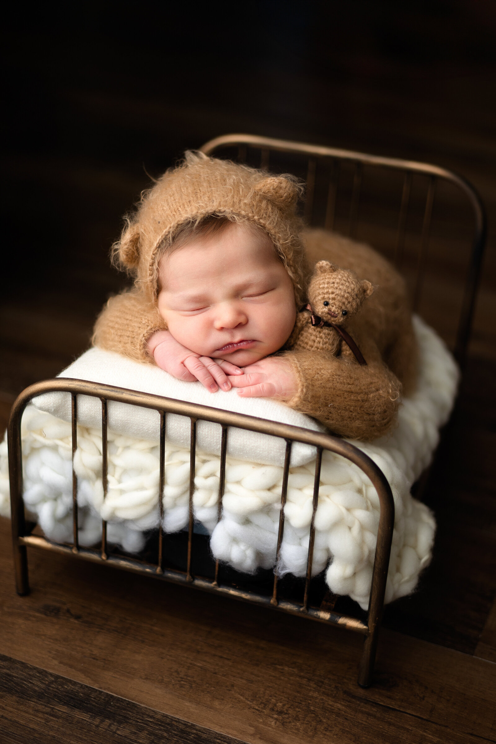 Newborn boy dressed in  bear outfit snuggled up on a small bed with his teddy bear