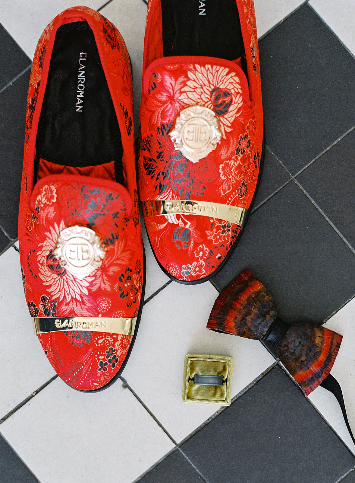 Groom's red shoes with gold and black details, red and black feather bowtie, gold ring box with groom's wedding band all on black and white diamond tile flooring. Photographed at Lowndes Grove wedding by wedding photographers in Charleston Amy Mulder Photography.