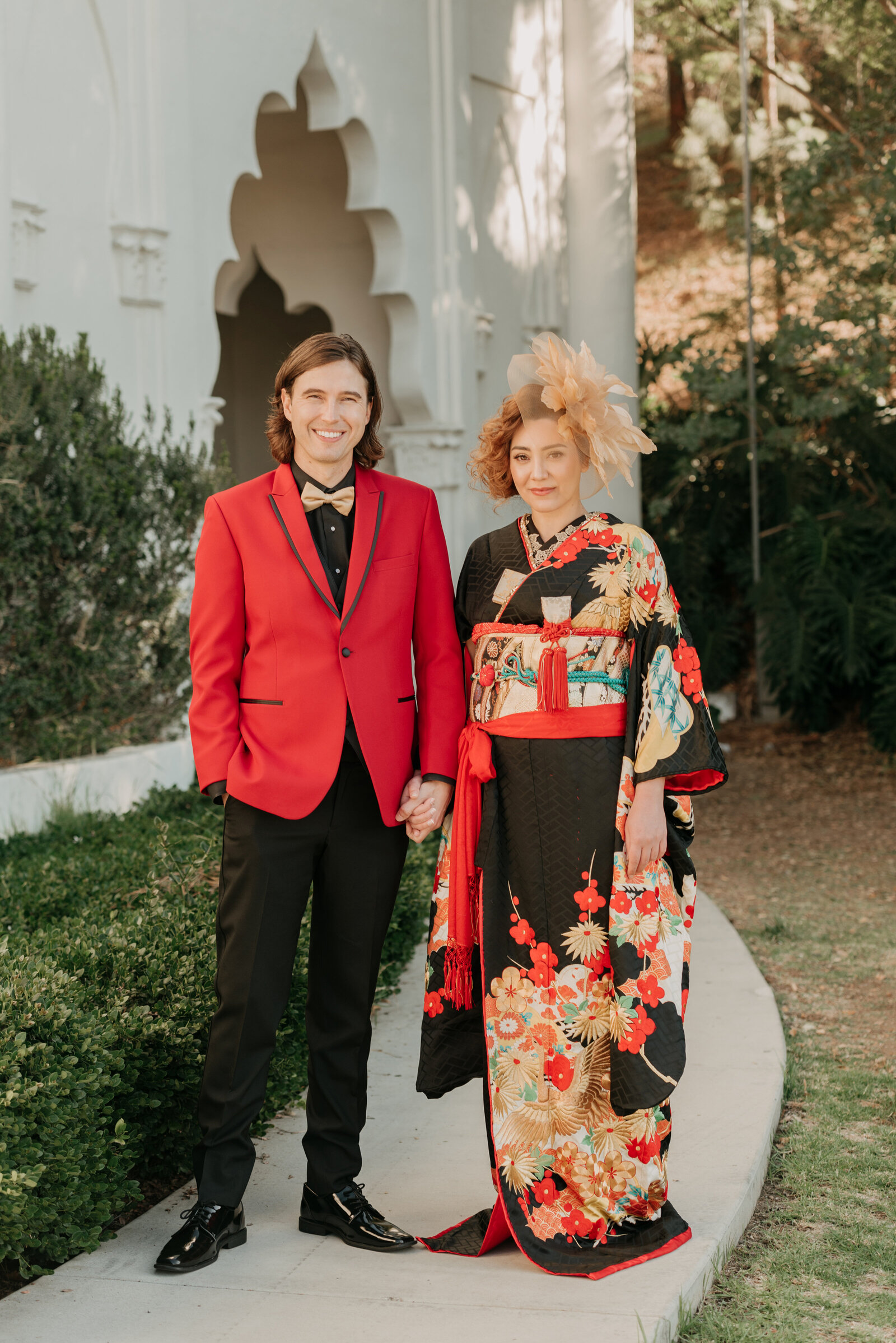 Bride in striking black and red wedding kimono rental with gold and floral design, standing hand in hand with groom in a vibrant red jacket at Brand Park, Los Angeles California | ブランドパークで赤黒打掛をレンタルした花嫁と赤ジャケット姿の新郎が手をつなぐウェディング撮影