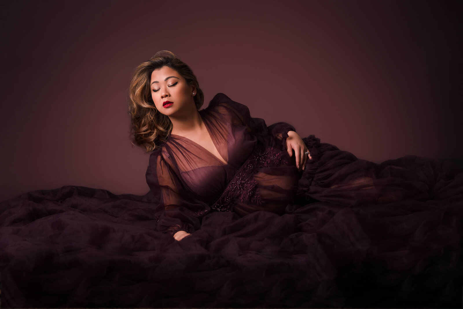 A pregnant woman reclines on flowing plum tulle fabric, her red lips and soft expression adding to the glamorous and regal atmosphere.