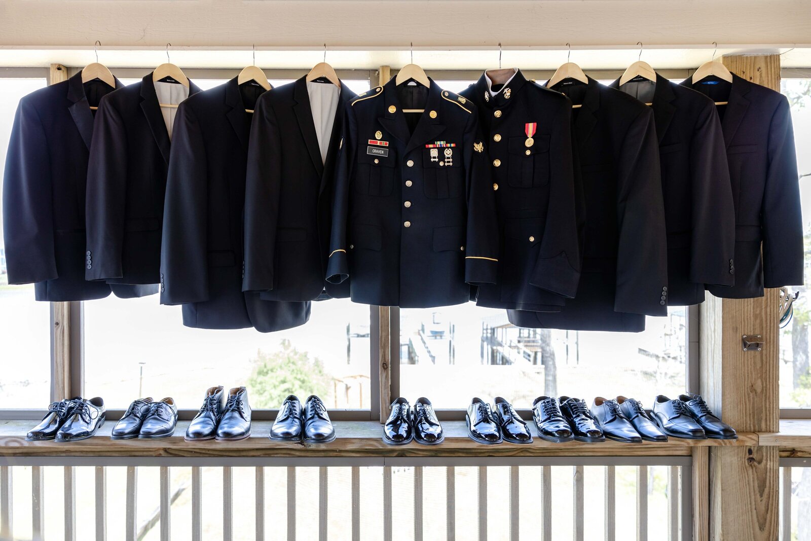 A lineup of groomsmen's suits and the Groom and Best Man's military uniforms hanging with polished dress shoes below, displayed at a coastal wedding venue in Oak Island, NC, blending tradition and elegance.