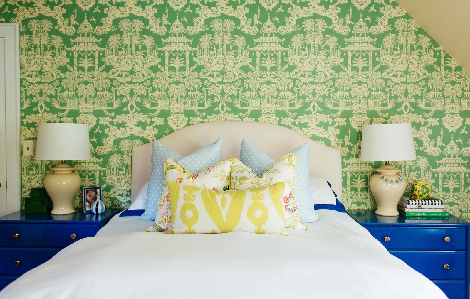 A white bed and blue end tables in front of a green wall papered wall.